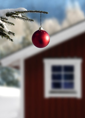 Ett rött hus med vita knutar i bakgrunden i ett snölandskap med en gren och julkula i förgrunden.
